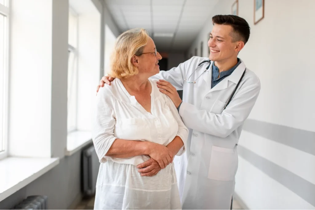 What Is Rheumatology? 11 Smiling doctor with a stethoscope comforting an elderly woman in a hospital hallway.