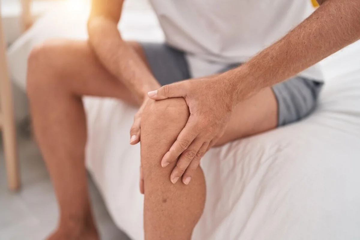 A man sitting on a bed, clutching his knee with both hands, indicating joint pain or discomfort.